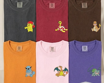 May include: Six Comfort Colors t-shirts in charcoal, maroon, brown, orange, light pink, and purple. Each shirt features a small embroidered character on the left chest, including Pikachu, Shuckle, Charmander, Squirtle, Charizard, and Bulbasaur.