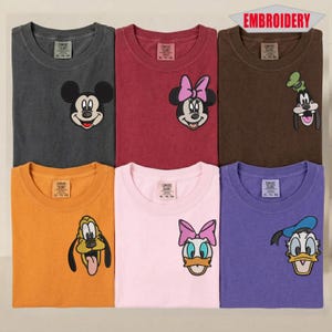 May include: Six colorful t-shirts with embroidered Disney character designs. The shirts are in various colors, including gray, maroon, brown, orange, pink, and purple. Each shirt features a different character, such as Mickey Mouse, Minnie Mouse, Goofy, Pluto, Daisy Duck, and Donald Duck.