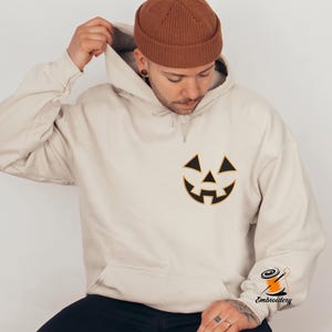 May include: A beige hooded sweatshirt featuring a black embroidered jack-o'-lantern face on the left chest. The hoodie includes a drawstring hood and a small orange spool of thread logo with the text Embroidery printed on the lower sleeve.