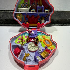 Può includere: Un set da gioco portatile in plastica rossa che si apre per rivelare l'interno di una casa delle bambole a più livelli. La sezione superiore presenta un bagno e una sala da pranzo, mentre quella inferiore raffigura un giardino con fontana.
