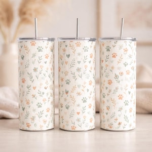 May include: Three stainless steel tumblers with lids and straws. The tumblers have a cream background with a pattern of paw prints, flowers, and leaves in pastel colours. The tumblers are cylindrical and appear to be approximately 20 cm tall.