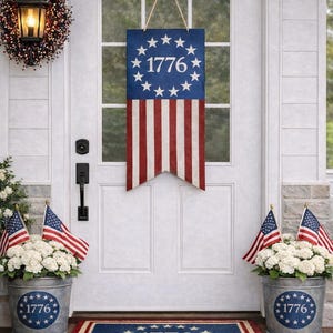 Op de afbeelding: Een patriottische voordeurdisplay met een blauw-rode banner met het jaartal "1776" en sterren, samen met bijpassende bloempotten en een deurmat. Amerikaanse vlaggen en witte bloemen dragen bij aan het feestelijke thema.