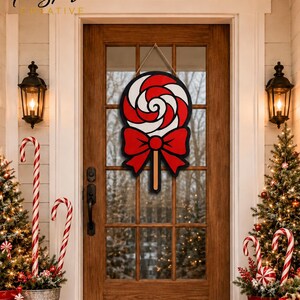 May include: A decorative wooden door hanger shaped like a round lollipop with red and white swirls, featuring a large red bow at the base of the candy head and a brown stick, suspended by a thin rope against a glass-paneled wooden door.