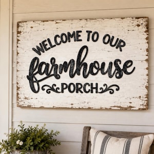 May include: A rectangular wooden sign with a distressed white finish featuring the black raised text "WELCOME TO OUR farmhouse PORCH" with decorative scrollwork accents at the bottom, mounted on a horizontal white paneled wall surface.