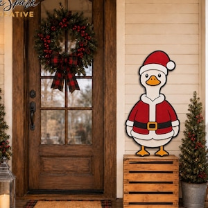 May include: A wooden cutout of a white goose wearing a red Santa suit, a red Santa hat with a white pom-pom, and a black belt with a gold buckle. The goose is standing on a light brown wooden crate next to a front door with a Christmas wreath.