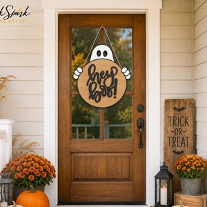 May include: A round wooden door sign featuring a white ghost illustration with black eyes and hands holding a circular plaque that displays the black text "hey boo!" in a script font. The sign hangs from a rope loop against a brown wooden door.
