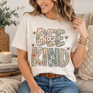 May include: Cream-colored t-shirt with the words "BEE KIND" in a floral and plaid design. The shirt features a brown belt and is paired with blue jeans. The design includes illustrations of bees.