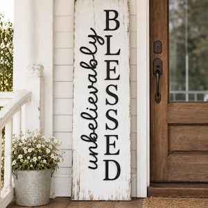 Puede incluir: Un letrero de porche rectangular y alto de madera con un acabado blanco desgastado. El letrero presenta el texto vertical negro en relieve "unbelievably BLESSED" escrito en una combinación de letra cursiva y de imprenta, apoyado sobre un suelo de madera.
