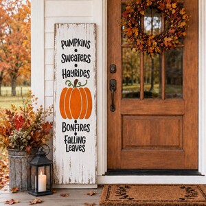Puede incluir: Un letrero de madera blanco, alto y rectangular, con acabado desgastado, que presenta el texto en negro PUMPKINS, SWEATERS, HAYRIDES, BONFIRES, FALLING LEAVES y una ilustración central de una calabaza naranja con tallo verde, en un porche.