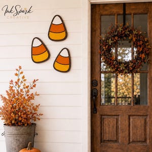 May include: A fall-themed porch decorated with candy corn wall art, a wreath, pumpkins, and fall foliage. The wooden door has glass panels. The porch has a galvanized metal bucket with orange branches and two pumpkins.