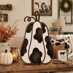 May include: A decorative wooden pumpkin cutout featuring a white base with dark brown, textured cow-print patches and a dark brown stem. The pumpkin has a layered design with visible depth and a smooth finish, standing upright as a tabletop ornament.