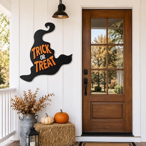 May include: A black witch hat-shaped wall sign featuring the orange text "TRICK OR TREAT" in a bold, sans-serif font. The sign is mounted on a white vertical paneled wall next to a wooden door, a metal milk can with foliage, and pumpkins.