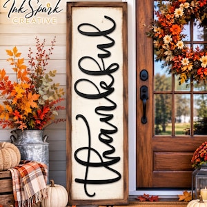 May include: A tall, rectangular wooden sign with a distressed white background and a dark brown frame. The word Thankful is written in large, black, cursive script lettering vertically down the center of the sign. The sign is placed on a porch.