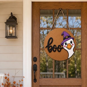 May include: A round wooden door sign featuring a white goose wearing a purple witch hat and the black script text "boo". The sign is suspended by a tan rope and displays a wood grain texture on the circular base, creating a seasonal decoration.