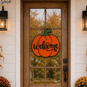 May include: A pumpkin-shaped wooden door sign featuring the word "welcome" in black script lettering. The sign is orange with a green stem and hangs from a thin rope. It is displayed on a brown wooden door with glass panels and a black handle.