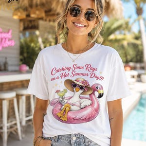 May include: A white short-sleeve t-shirt featuring a graphic of a white duck wearing a sun hat and heart-shaped sunglasses while floating in a pink flamingo pool float, holding a green cocktail, with the text Catching Some Rays on a Hot Summer Day.