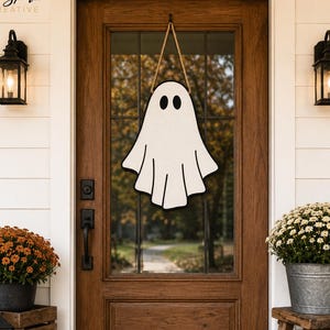 May include: A white ghost-shaped door hanger with black eyes and mouth, suspended by a tan rope. The ghost features a simple, flat design with a scalloped bottom edge, hanging centred on a wooden door with a glass panel and black hardware.