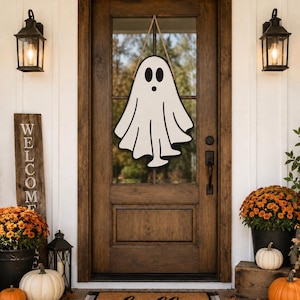 May include: A white ghost-shaped wooden door hanger with black eyes and a black mouth, suspended by a tan rope. The ghost features a simple, cartoon-like design with a wavy bottom edge, hanging centred on a dark brown wooden door with glass panels.