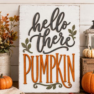 Puede incluir: Un letrero rectangular de madera blanca con acabado desgastado que presenta el texto en relieve negro "hello there" y el gran texto en relieve naranja "PUMPKIN". El letrero incluye pequeños detalles de hojas verdes.