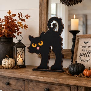 May include: A black wooden cat cutout with an arched back, a curled tail, and bright orange eyes. The cat has a small pink nose and mouth, and the edges of the black silhouette are distressed with a copper-colored finish, standing on a base.