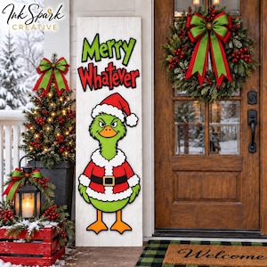 May include: A tall white wooden porch sign featuring the green text Merry and red text Whatever above a cartoon green duck wearing a red and white Santa suit and hat. The sign is displayed next to a Christmas tree, a lantern, and a wreath.