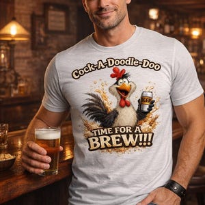 Puede incluir: Camiseta gris claro con el texto "Cock-A-Doodle-Doo" y un gallo de dibujos animados sosteniendo una lata de cerveza. La camiseta también dice "TIME FOR A BREW!!!". Una persona sostiene un vaso de cerveza.