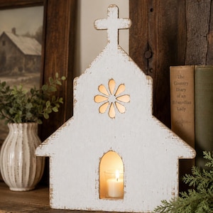 May include: A white, distressed wooden church-shaped candle holder featuring a cross at the peak, a flower-shaped cutout near the top, and an arched opening at the base containing a small glass votive holder with a lit white candle inside.