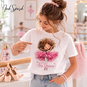 May include: White t-shirt with a ballerina graphic in a pink tutu and the text "Tiny Dancer, Big Dreams." The image also shows a pink water bottle and ballet shoes.