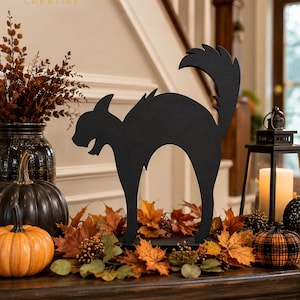 May include: A black wooden cutout of an arched-back cat with a raised tail. The silhouette features a pointed ear and an open mouth. The cat stands on a flat base, positioned among orange and brown autumn leaves, pumpkins, and a black lantern.