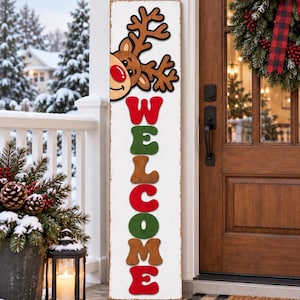 May include: A tall, rectangular white wooden sign featuring a cartoon reindeer head with a red nose at the top and the word WELCOME written vertically in alternating red, green, and brown block letters. The sign has a distressed finish.