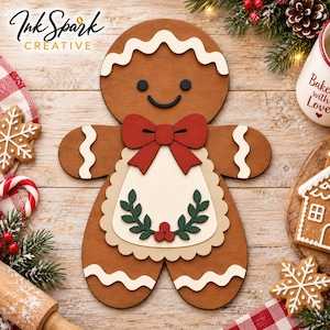 May include: A brown wooden gingerbread person cutout featuring white wavy icing details, a red bow, and a cream-colored apron decorated with a green leaf and red berry wreath. The figure has black circular eyes and a curved black mouth.