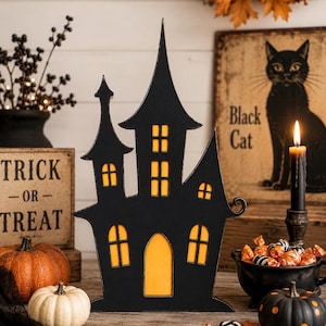 May include: A black wooden haunted house silhouette decoration featuring cut-out windows and a door backed with glowing orange light. The structure has a tall central spire and two smaller side towers, creating a flat, decorative Halloween-themed display piece.