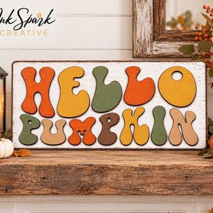 Peut inclure: Un panneau rectangulaire en bois avec un fond blanc vieilli portant l'inscription HELLO PUMPKIN en grandes lettres arrondies et multicolores. Les lettres sont déclinées dans des tons orange, jaune, vert olive et marron avec une police ondulée.