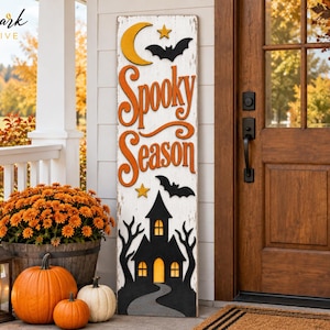 Pode incluir: Uma placa de madeira alta e retangular para varanda com acabamento branco desgastado. Apresenta uma lua crescente amarela, estrelas, morcegos pretos e o texto laranja "Spooky Season" sobre uma silhueta preta de uma casa assombrada.