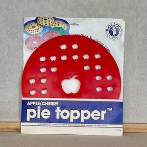 May include: A red circular plastic pie topper with multiple apple-shaped cutouts, packaged on a white and blue cardboard backing. The packaging features the text APPLE/CHERRY pie topper, the Rowoco logo, and images of various baked pies.