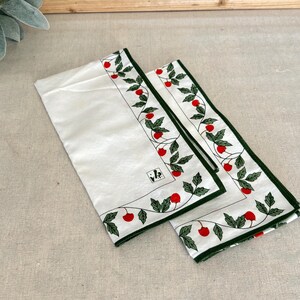 May include: Two white rectangular napkins featuring a decorative border of green leaves and red berries. The fabric has a dark green hem and a small square label with black text printed on the surface of the larger napkin.