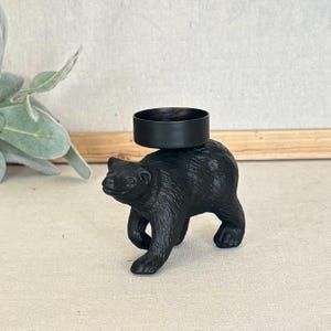 May include: A matte black bear figurine serving as a candle holder. The bear is depicted in a walking stance with textured fur details. A circular, black metal cup is positioned on the back of the bear to hold a standard tea light candle.