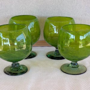 May include: Four green glass goblets with rounded bowls and short stems. The glasses have a vibrant, transparent green color and are arranged on a white surface. The goblets are ideal for serving beverages.