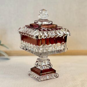 May include: A vintage, ruby-colored glass candy dish with a lid. The dish has a square base and a pedestal, with a decorative, scalloped edge around the rim. The lid is topped with a small, ornate finial. The glass is clear with ruby accents.