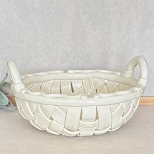 May include: A cream-colored ceramic basket featuring a woven design with a glossy finish. The basket has a rounded body, a thick rim, and two curved handles on opposite sides. The surface texture mimics the appearance of woven strips.