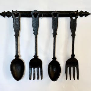 May include: A black cast iron kitchen utensil rack with four hanging pieces: two spoons and two forks. The rack has decorative arrow-shaped ends. The utensils have ornate handles and are suspended from the rack.