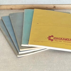 May include: A stack of five softcover notebooks arranged in a fan shape. The top notebook is yellow with red text reading CHANDA and VALLABH VIDYANAGAR, while the others are shades of blue and gray. Each cover features horizontal lines for writing.