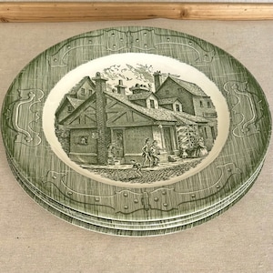 May include: A stack of round ceramic plates featuring a cream-colored center with a detailed black ink illustration of a historic village scene including buildings and figures. The wide green border has a textured finish and decorative hinge motifs.