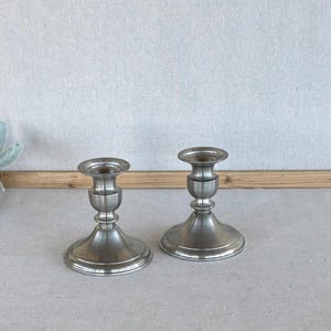 May include: Two matching silver-toned metal candle holders with a smooth, polished finish. Each piece features a wide, circular base, a slender central stem with decorative ring detailing, and a flared top cup designed to hold a single candle.