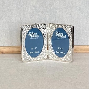 May include: A silver-plated double photo frame with an intricate embossed floral pattern. Each side features an oval opening for a 3 inch by 4 inch photograph. The blue inserts are printed with the text Silver Plated Photo Frame 3 inch x 4 inch 8cm x 10cm.