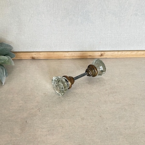 May include: A door knob set featuring two clear, faceted glass knobs connected by a central metal spindle. Each glass knob is attached to a circular, aged brass-toned metal base, providing a contrast between the transparent glass and the metallic components.