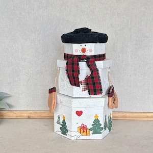 May include: A decorative snowman figure constructed from three stacked white hexagonal boxes. The snowman wears a black knit hat and a red and black plaid scarf. The base box features painted green pine trees, a red heart, and a yellow gift box.
