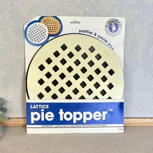 May include: A round, cream-colored plastic lattice pie topper in its original packaging. The packaging is white and blue with the text 'LATTICE pie topper', 'prettier & juicier pies', and the Rowoco logo. It includes a small circular image of a pie.
