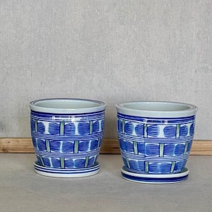 May include: Two cylindrical ceramic planters with matching saucers, featuring a white base decorated with horizontal blue bands and a repeating pattern of blue brushstroke motifs separated by thin vertical green lines.