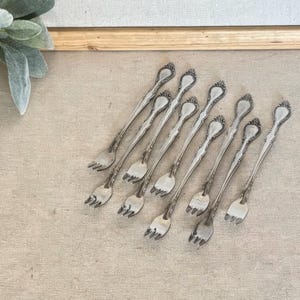 May include: A set of eight silver-toned metal cocktail forks arranged in a diagonal pattern. Each fork features a long, slender handle with an ornate, embossed floral detail at the top and three short, pointed tines at the base.